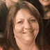 Smiling woman with straight, shoulder-length brown hair in a reddish-brown top, foregrounded with others blurred behind her.
