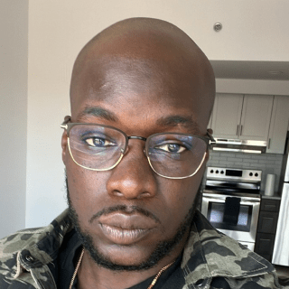 A man with glasses and a trimmed beard wearing a camo jacket and gold necklace stands in a modern kitchen with white cabinets.