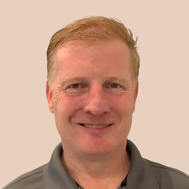 Smiling man with short, light reddish-blond hair in a gray collared shirt stands before a beige background, representing Field Sales Software.