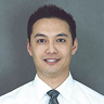 A smiling man with short dark hair in a white shirt and gray tie stands before a plain gray background, ready to discuss route planning software.