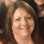 Smiling woman with straight, shoulder-length brown hair in a reddish-brown top, foregrounded with others blurred behind her.