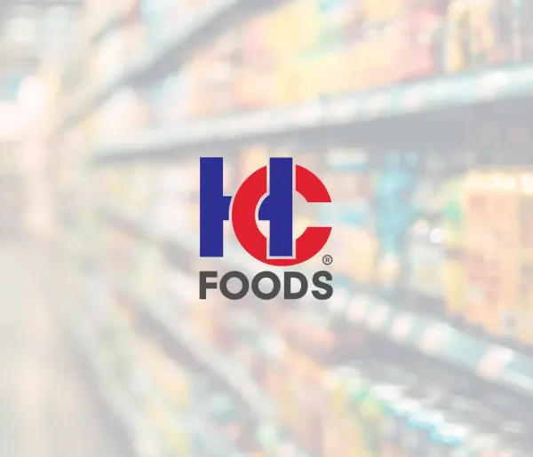 Centered against a blurred grocery store aisle, the HC Foods logo stands out in blue and red amid shelves of assorted products.