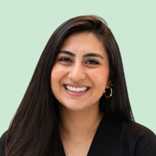 Smiling young woman with long dark hair, gold hoop earrings, and a black top stands in front of a light green background.